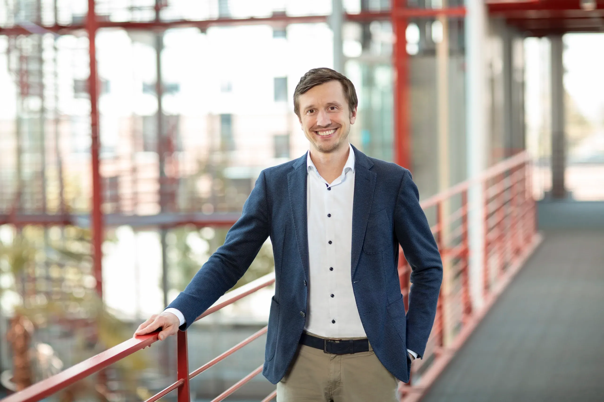 Bereichsleiter Cornelius Kiermarsch im Business-Outfit lehnt an einem Geländer in einem modernen Bürogebäude mit viel Glas und roten Akzenten.
