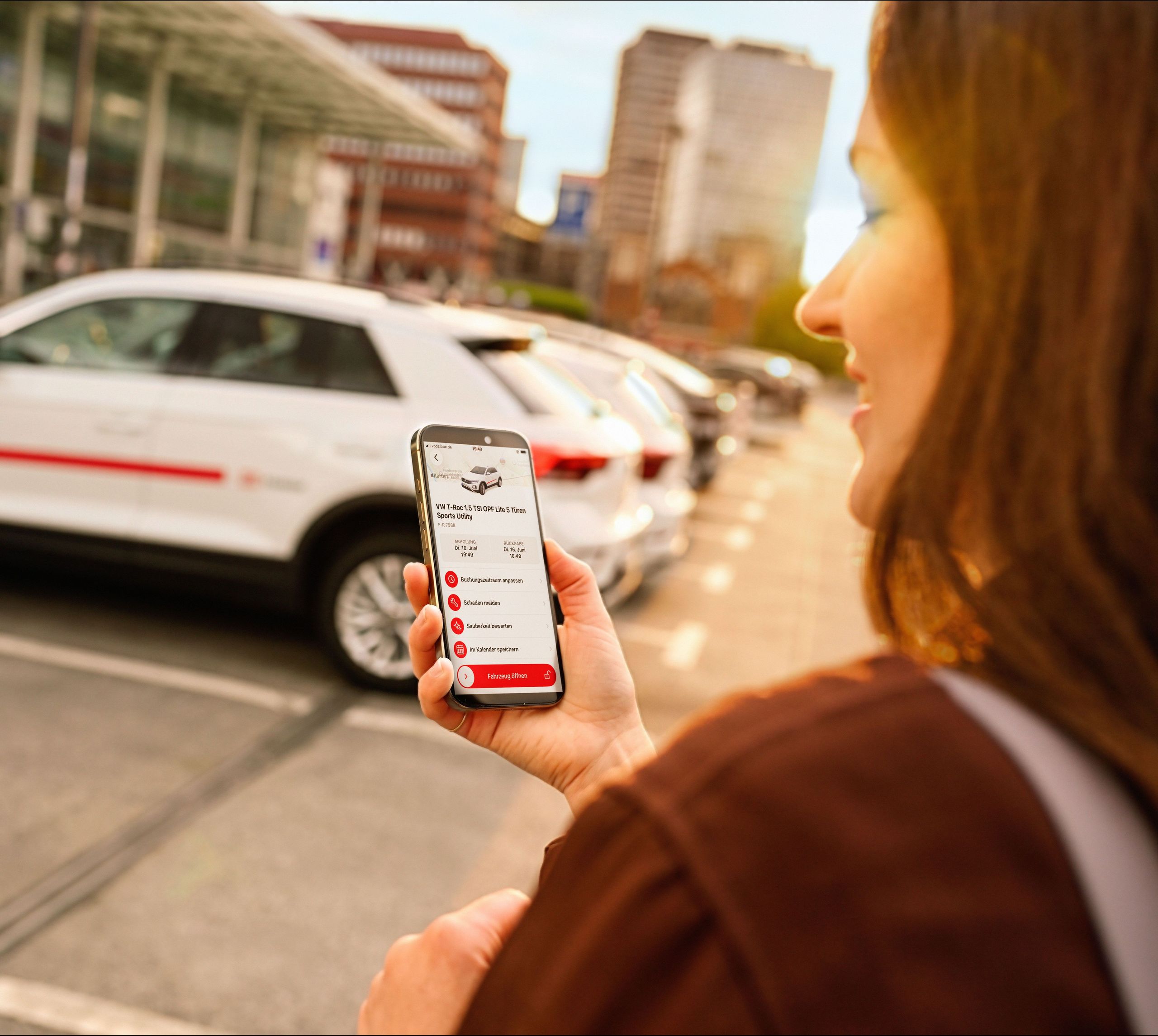A Flinkster customer standing in front of a white Flinkster car, looking at their smartphone with the Flinkster app open.