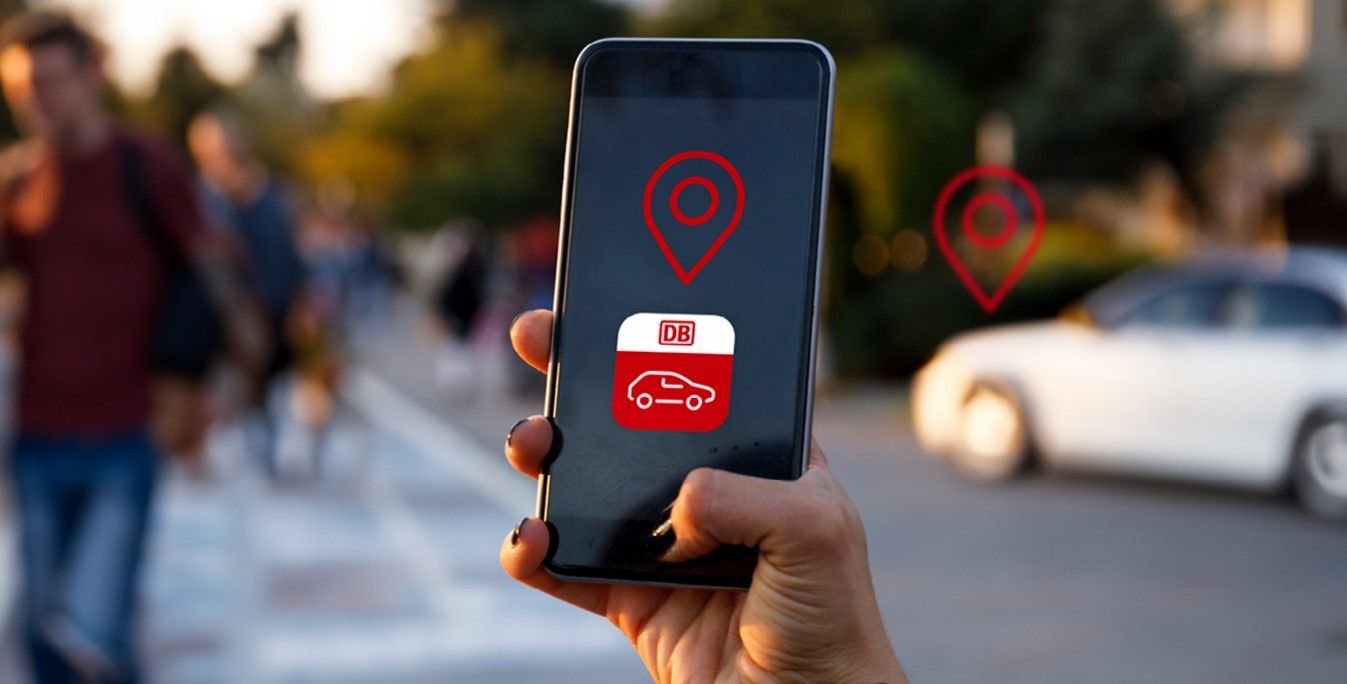 Hand holding a smartphone with the Flinkster app open, people and a Flinkster car can be seen blurred in the background.
