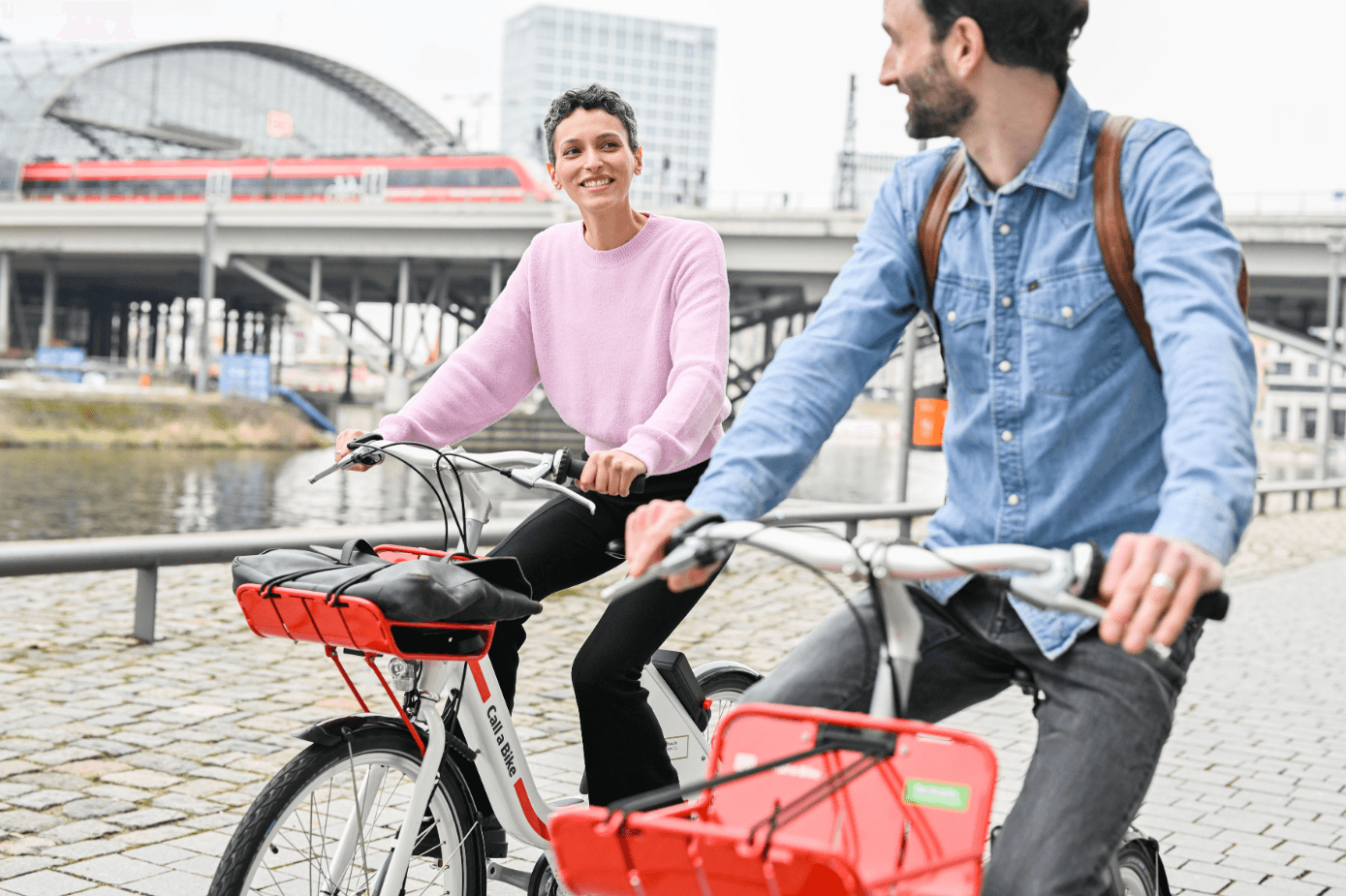 Two people on Call a Bike bicycles ride along a river with Berlin Central Station and a train in the background.