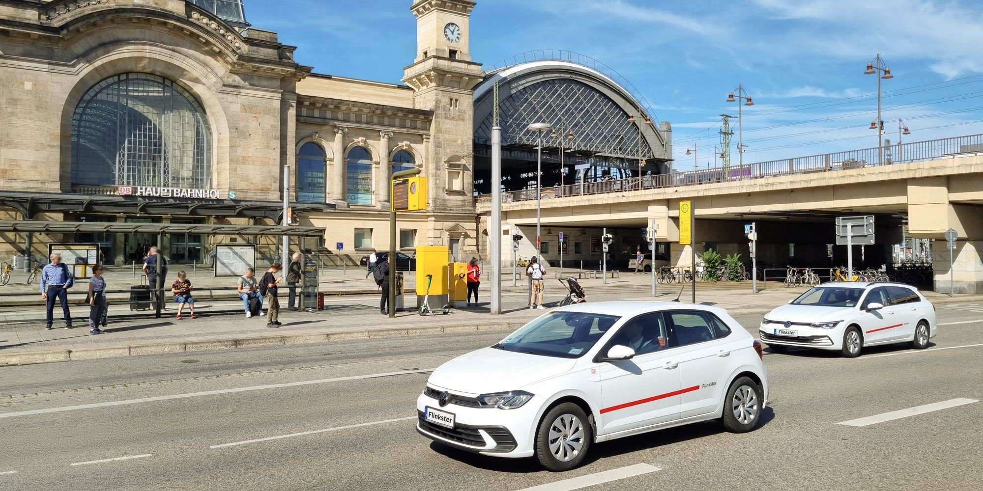 Two white Flinkster vehicles, a VW Polo and a VW Golf Variant, drive past in front of the main station. In the background, people are waiting at a bus stop.