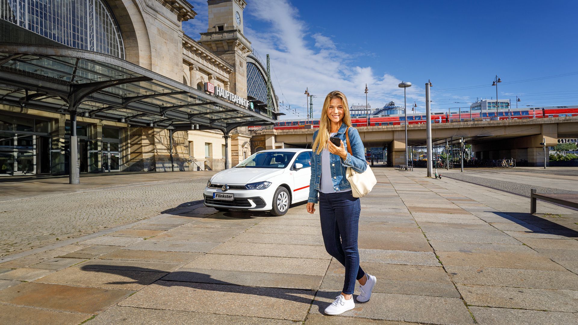 Woman with smartphone and shoulder bag at the station with Flinkster vehicle in the background.
