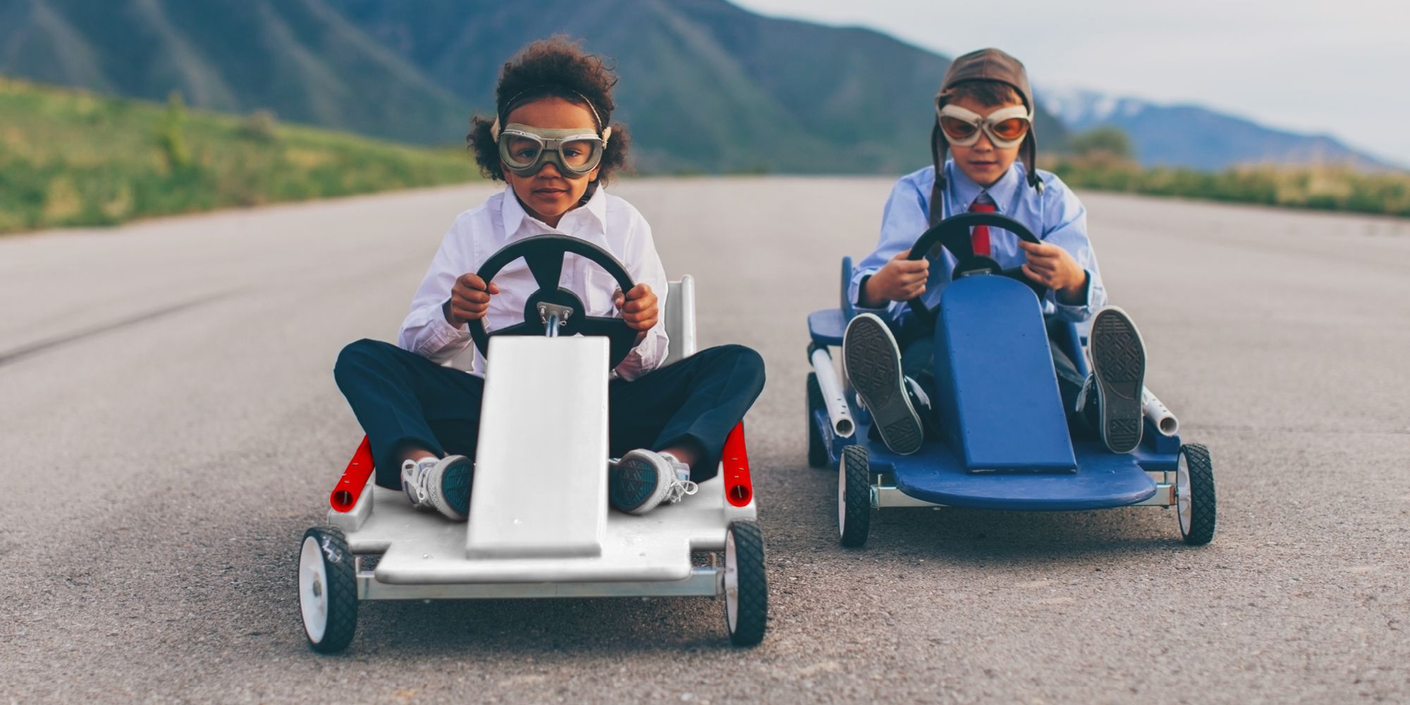Two children wearing safety goggles and driving on a country road in home-made soapboxes. The soapboxes are white and blue, and mountains can be seen in the background.