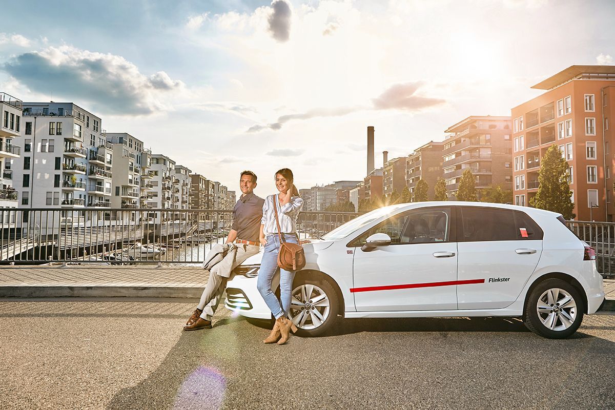 A man and a woman stand relaxed by a white Flinkster car on a bridge in a modern city. Residential buildings and water can be seen in the background.