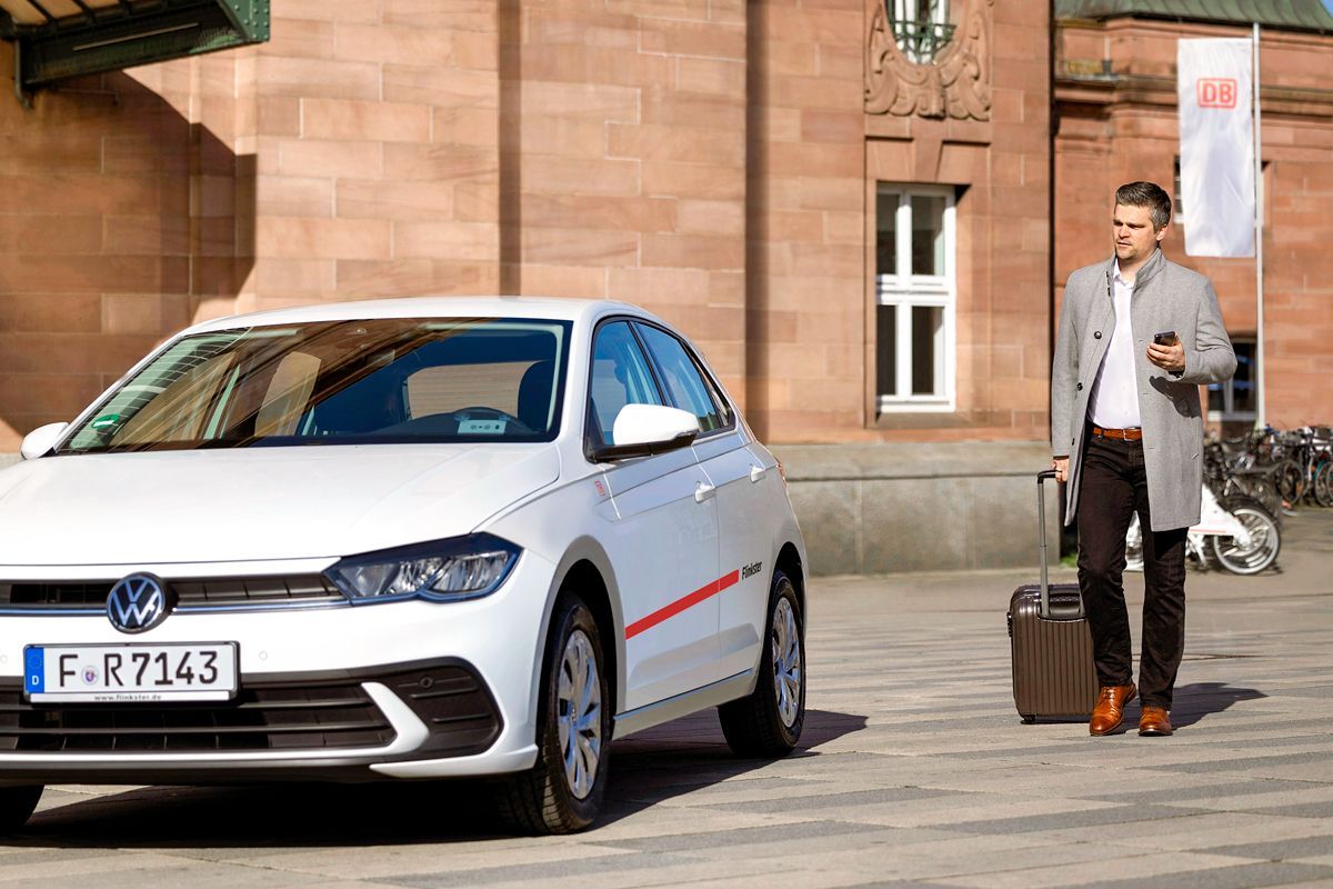 A man with a coat and rolling suitcase walks towards a white Flinkster car parked in front of a historic passenger building with the DB logo.