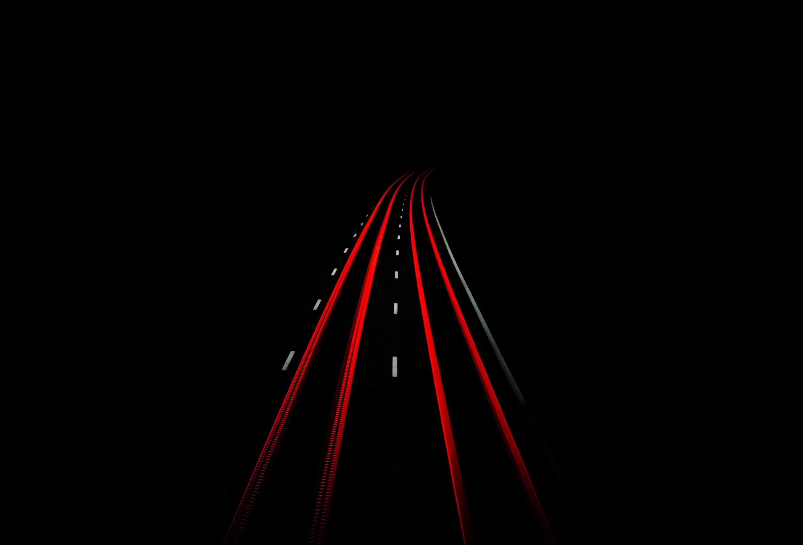 Eine nächtliche Langzeitbelichtung einer Straße zeigt rote Lichtspuren von Fahrzeugen, die sich entlang der Fahrbahn erstrecken und in der Dunkelheit verschwinden. Die Straße selbst ist nur schwach beleuchtet, wodurch die Lichtspuren besonders hervortreten.