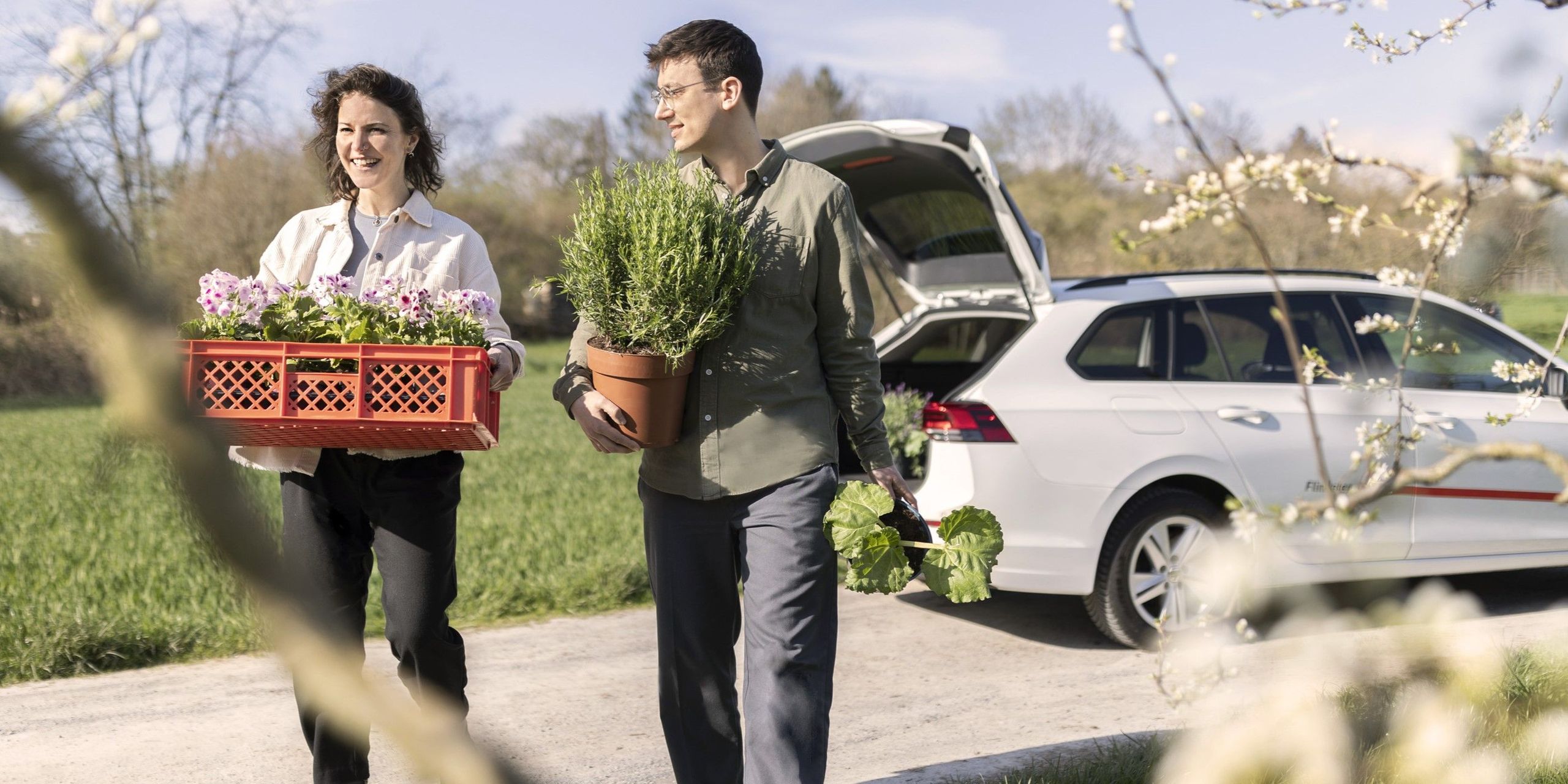Ein Mann und eine Frau tragen Pflanzen und Setzlinge. Im Hintergrund steht ein Flinkster-Auto mit geöffnetem Kofferraum.