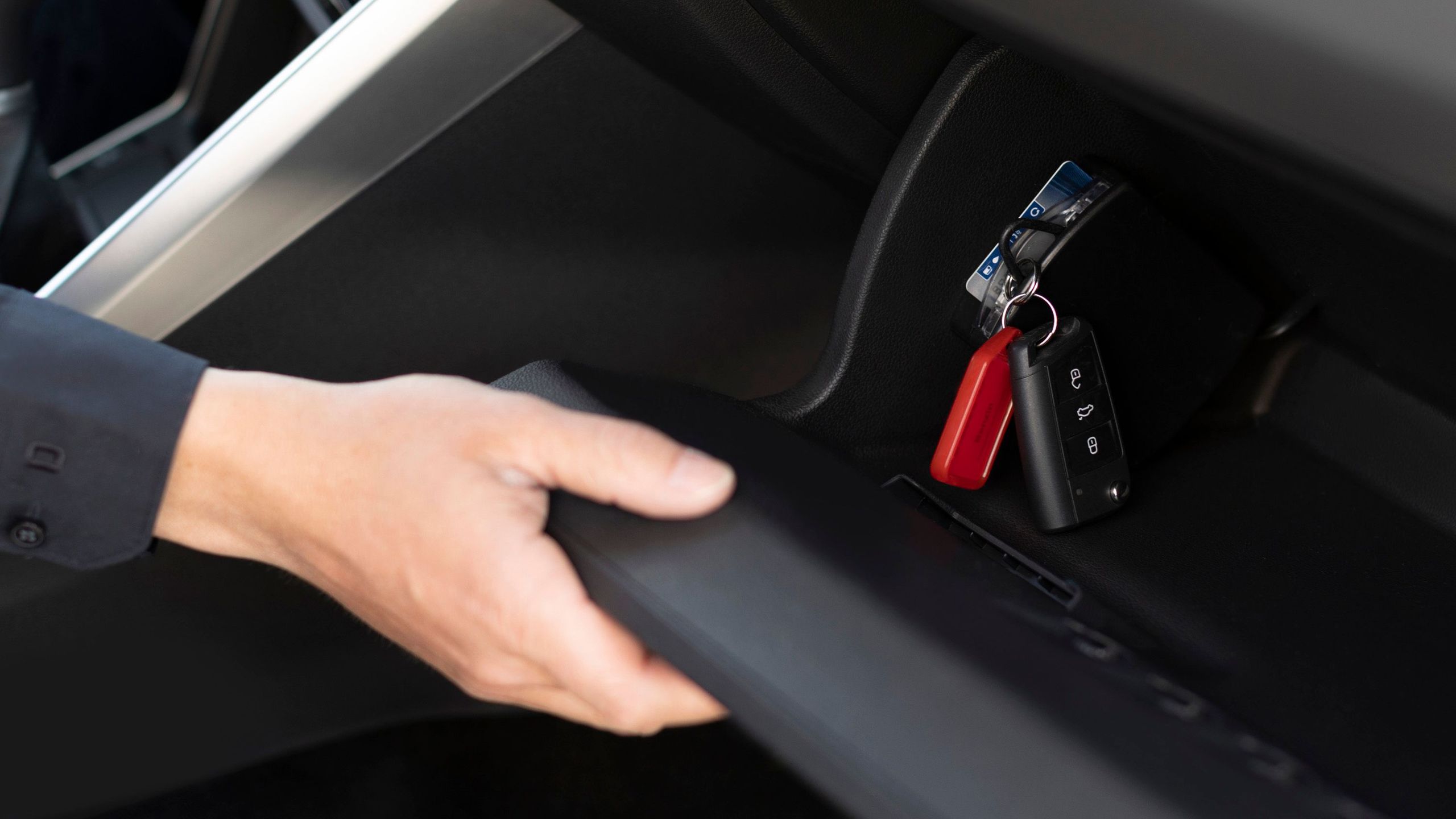 Interior of a car with the footwell flap on the passenger seat opened by one hand. The vehicle key is inserted into a carsharing module inside the compartment.