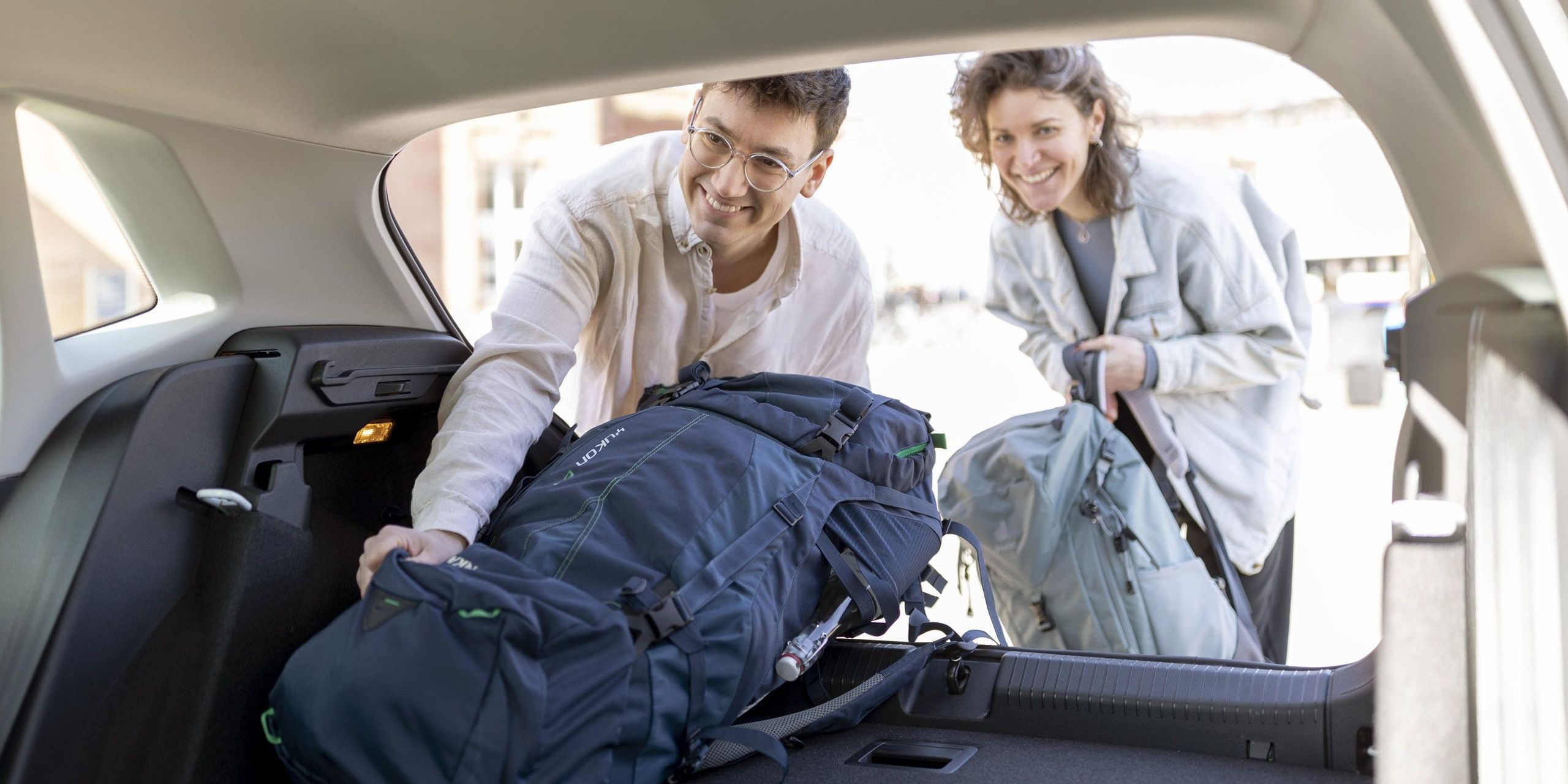 Ein Mann und eine Frau laden lächelnd ihre Reiserucksäcke in den Kofferraum eines Flinkster-Autos.