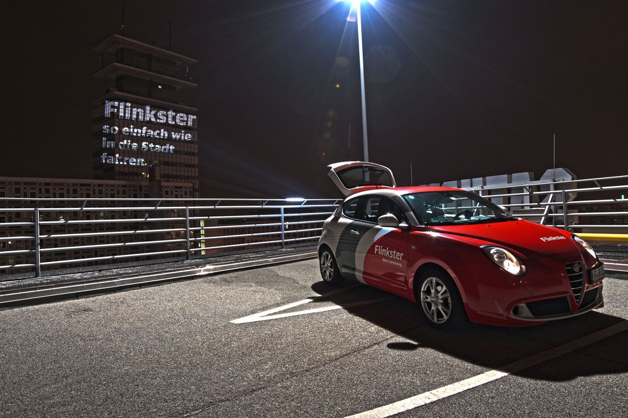 Flinkster car with open trunk at night in a parking lot, projecting the message 'Flinkster as easy as driving into the city' onto a dark house wall.