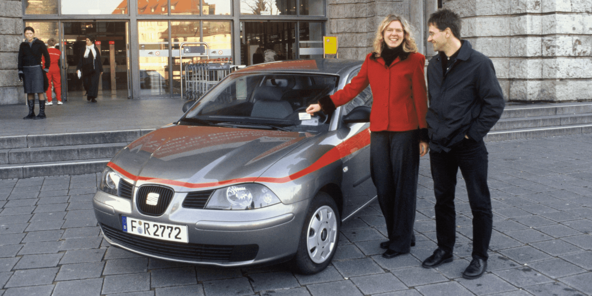 Two people are standing next to a gray DB carsharing car in front of a passenger building. copyright DB AG / Weber