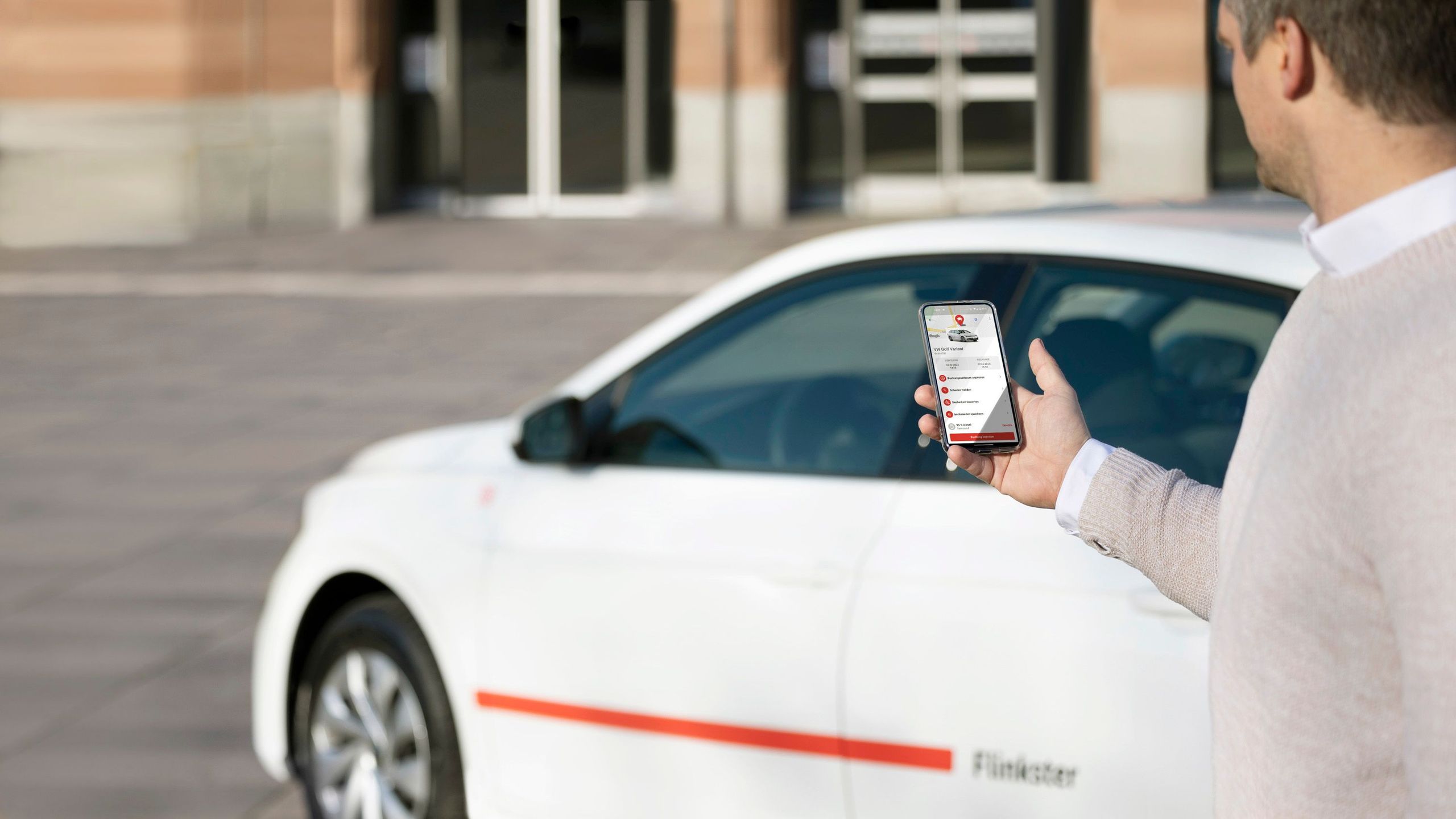 Flinkster-Kunde vor einem weißen Flinkster-Auto, schaut auf sein Smartphone, auf dem die Flinkster-App geöffnet ist.