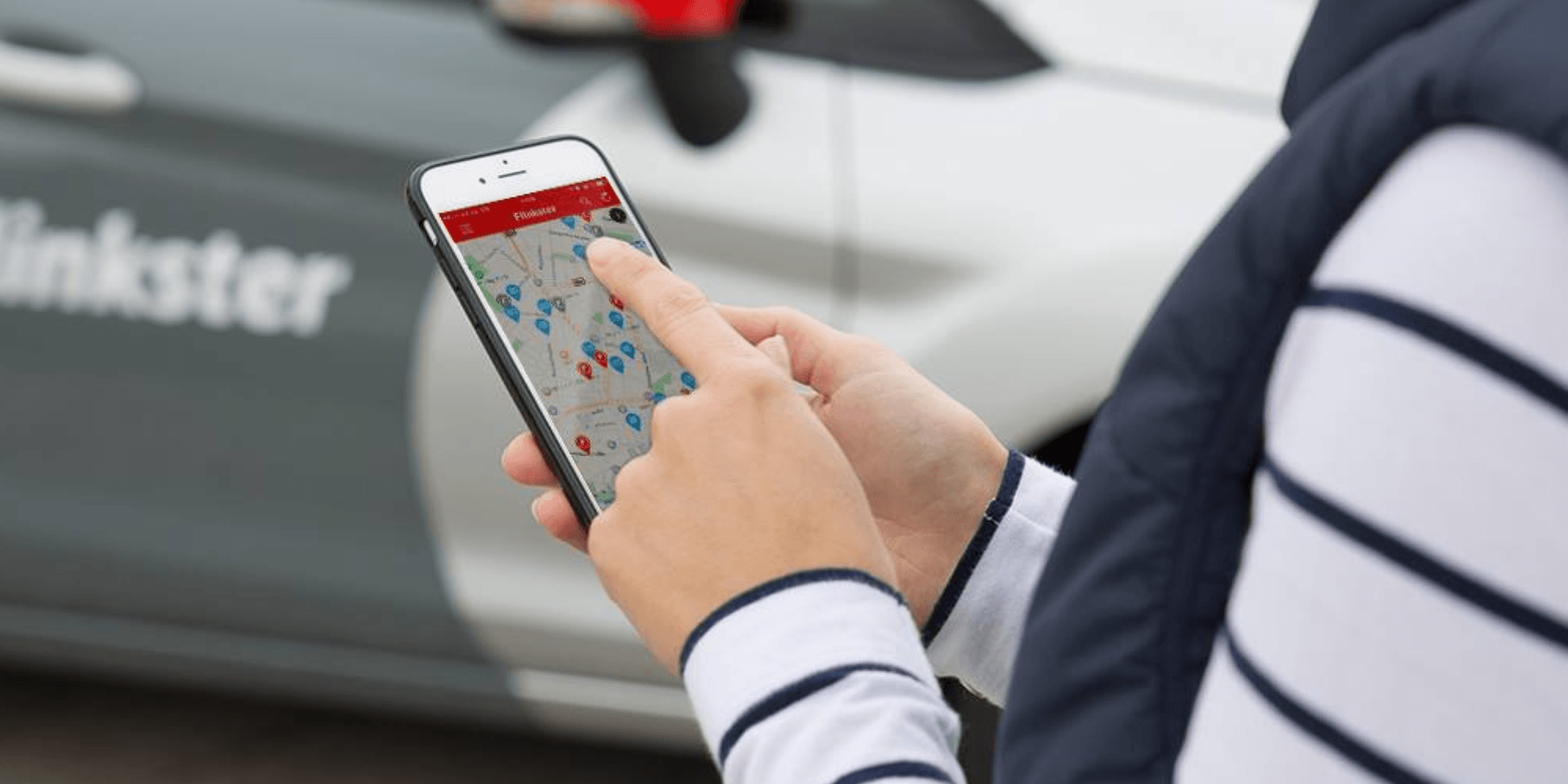 Person benutzt ein Smartphone, auf dem die Flinkster-App geöffnet ist, im Hintergrund ein Flinkster-Auto.