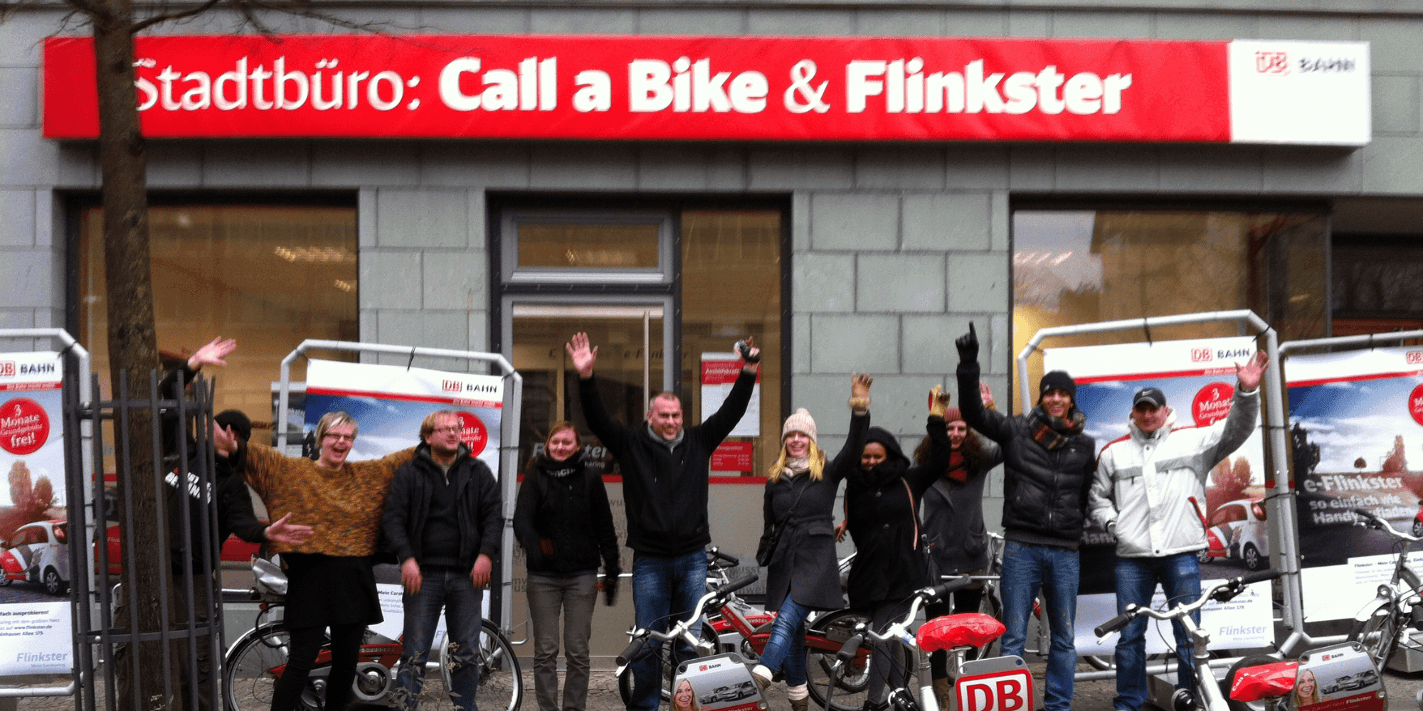 Fröhliche Gruppe von Menschen posiert vor einem Stadtbüro mit dem Schild „Call a Bike & Flinkster“, jubelt und steht neben Fahrrädern und Werbematerial für Fahrrad- und Carsharingdienste.