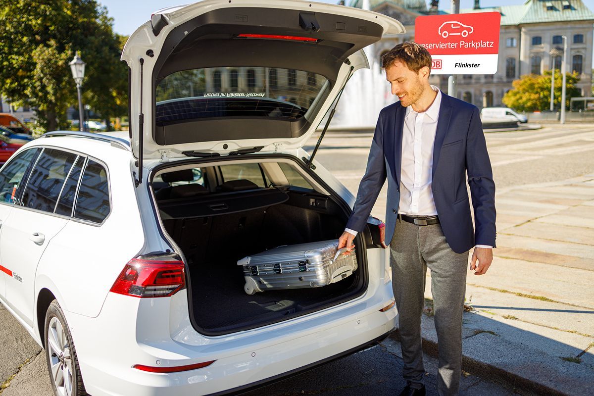 A man in business attire loads a silver suitcase into the trunk of a white Flinkster car parked in a parking lot with a Flinkster parking sign.