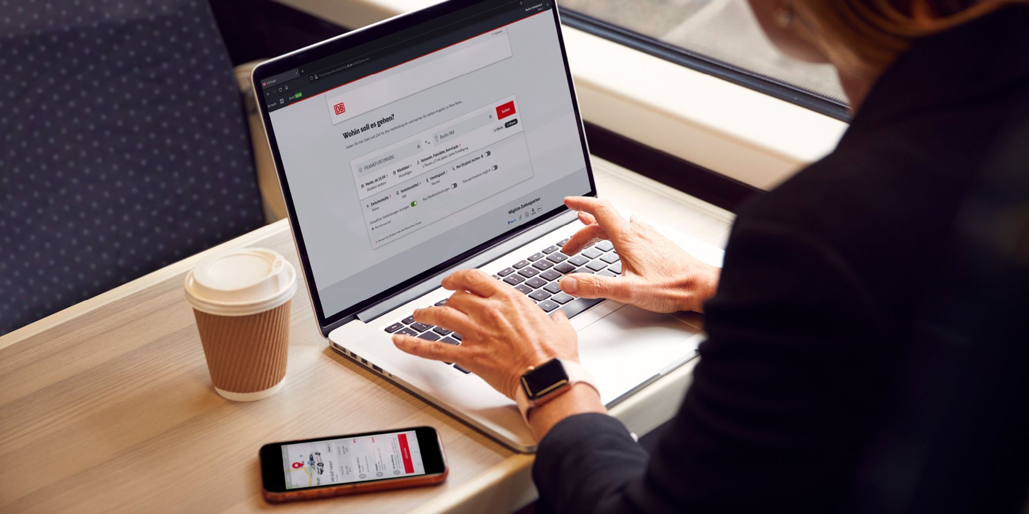 A woman is sitting on the train, working on a laptop with DB Navigator open. Next to her is a coffee mug and a smartphone with the Flinkster app open.